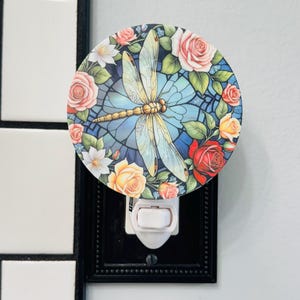 May include: A decorative night light featuring a dragonfly design. The round light has a blue background with a dragonfly in the center, surrounded by roses in shades of pink, yellow, and red. The night light is set in a black frame.