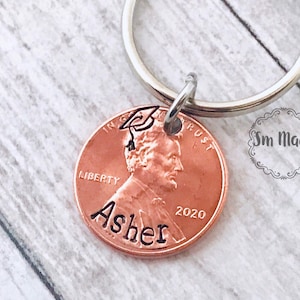 May include: A copper penny keychain with a graduation cap engraved on it. The penny has the words "Liberty", "In God We Trust", and "2020" engraved on it. The name "Asher" is engraved on the bottom of the penny.