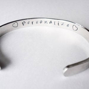 May include: A silver bangle bracelet with the words "Personalize" engraved on it, with a heart on each side of the text.