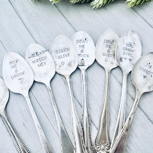 May include: A collection of vintage silver spoons with stamped phrases. The spoons feature various sayings like "Just let me die," "Pour me a drink," and "I'm thirsty." The spoons are arranged on a light-colored wooden surface.