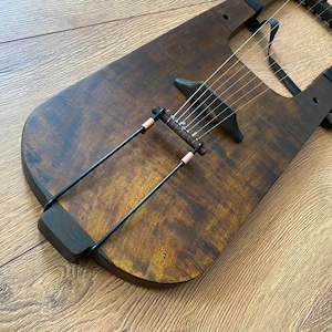 May include: A wooden lyre with a dark brown stain. The lyre has 8 strings and a black metal tuning peg. The lyre is on a wooden surface.