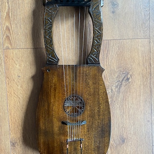 Puede incluir: Una lira de madera con acabado marrón oscuro. El instrumento presenta detalles tallados en el marco y una boca con una rejilla metálica decorativa. Las cuerdas son visibles, tensadas a través del cuerpo del instrumento.
