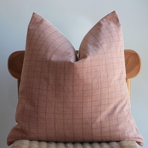 Puede incluir: Un cojín cuadrado grande con un patrón de cuadrícula en tonos rosa empolvado y marrón. El cojín está colocado sobre una silla de madera con un asiento tejido. La textura y el color del cojín crean una estética cálida y acogedora.