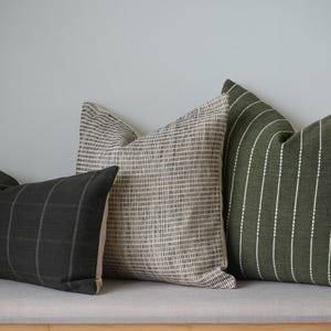 Puede incluir: Tres almohadas decorativas con diferentes patrones. La almohada de la izquierda es negra con finas rayas blancas. La almohada del medio es beige con un sutil patrón marrón. La almohada de la derecha es verde con rayas blancas.