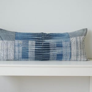 May include: A rectangular lumbar cushion featuring a patchwork design of various blue and grey fabric panels. The textile displays a mix of solid denim-like textures, vertical stripes, and intricate white geometric patterns across the surface.
