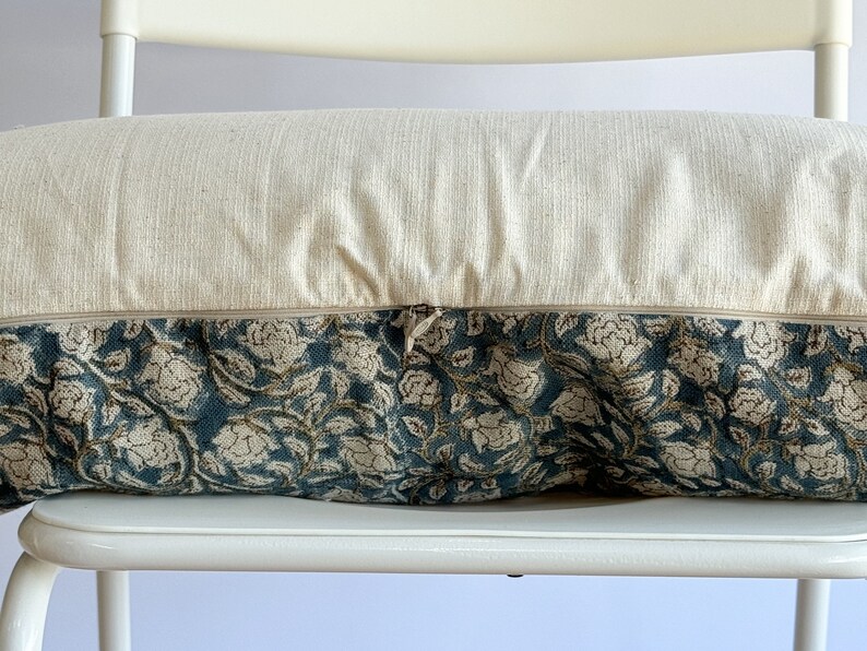 May include: A cream-colored pillow with a zipper closure and a floral patterned fabric. The pillow is sitting on a white chair.