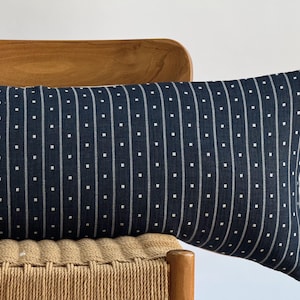 May include: A long, rectangular navy blue pillow with a white dotted stripe pattern. The pillow is resting on a wooden chair with a woven seat, creating a contrast in textures and colors.