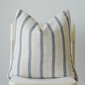 Könnte beinhalten: Ein dekoratives Kissen mit vertikalen blauen Streifen auf cremefarbenem Hintergrund. Es liegt auf einem weißen Stuhl. Das schlichte Design passt zu verschiedenen Einrichtungsstilen. Ideal für Sofa oder Bett.