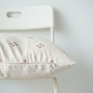 May include: A white chair with a beige throw pillow with a brown geometric pattern. The pillow has a zipper closure.