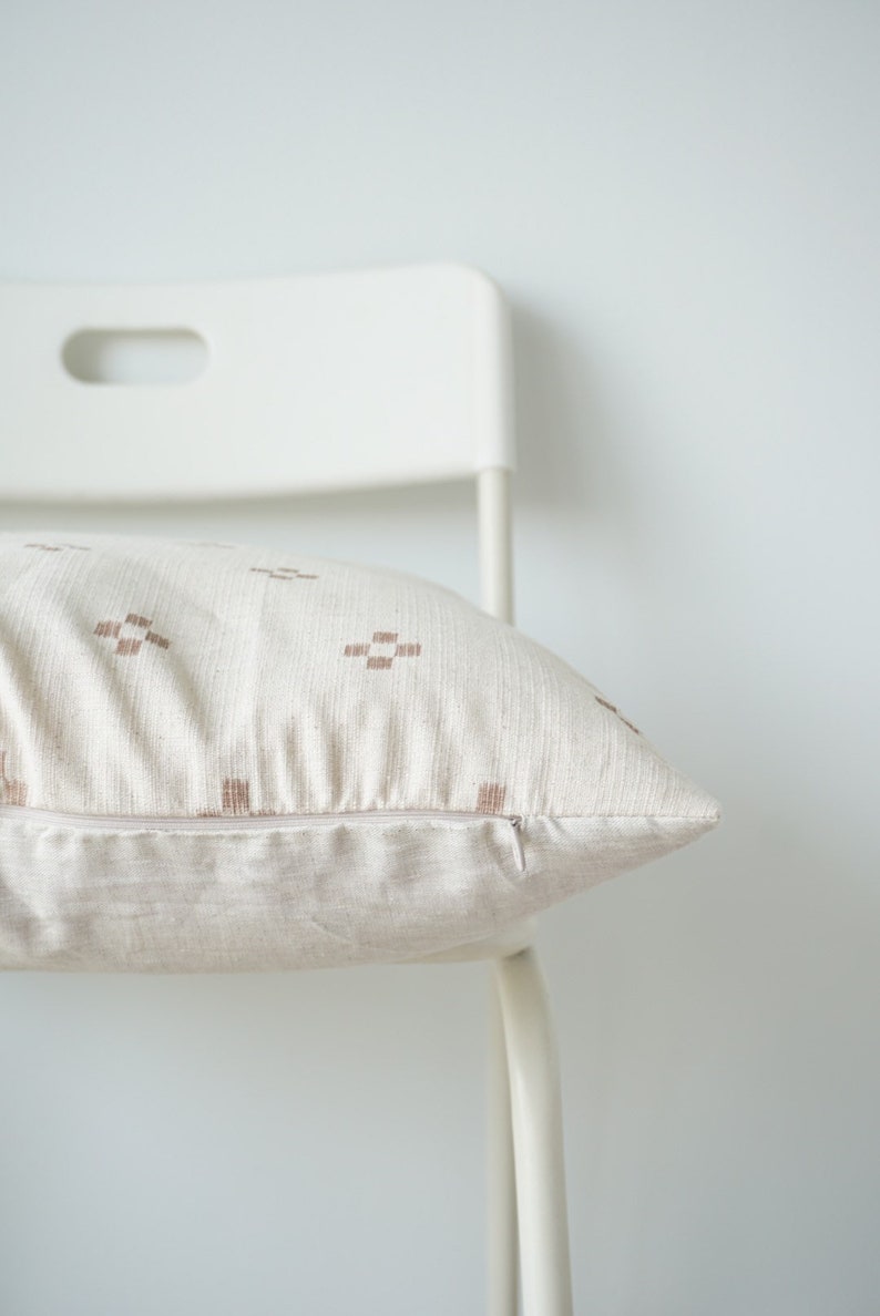 May include: A white chair with a beige throw pillow with a brown geometric pattern. The pillow has a zipper closure.