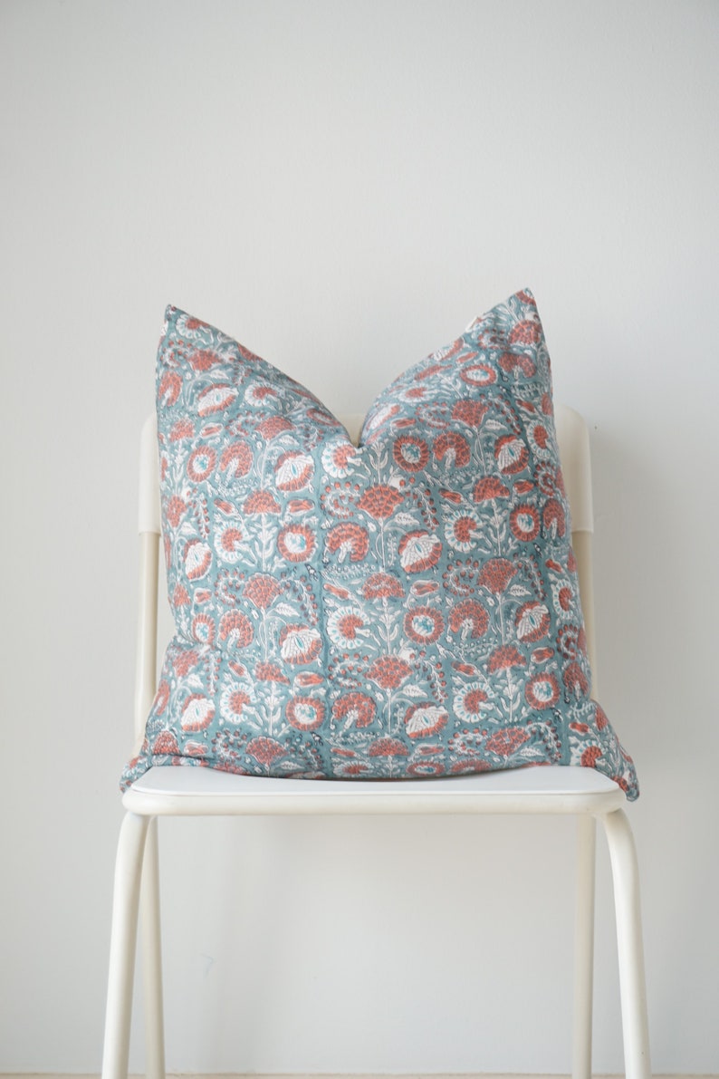 Puede incluir: Una almohada con estampado floral de color azul verdoso y coral sobre una silla blanca.