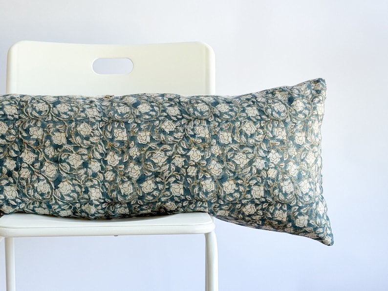 May include: A teal blue and white floral patterned lumbar pillow resting on a white chair.