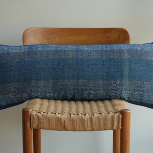 Puede incluir: Un cojín rectangular azul con textura tejida, apoyado sobre una silla tejida. El cojín tiene un patrón de rayas en tonos azules y beige. La silla tiene un marco de madera y un asiento tejido.