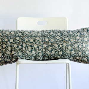 May include: A long, rectangular pillow with a teal background and a white floral pattern. The pillow is sitting on a white chair.