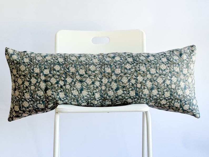 May include: A long, rectangular pillow with a teal background and a white floral pattern. The pillow is sitting on a white chair.