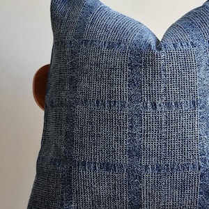 May include: A square, indigo blue pillow with a textured, woven pattern. The pillow's fabric has a grid-like design, creating a visual texture. The pillow is resting on a wooden chair.