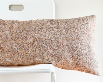 Puede incluir: Una almohada larga y rectangular con estampado floral. La almohada está hecha de una tela marrón claro con un patrón floral blanco. La almohada está sentada en una silla blanca.