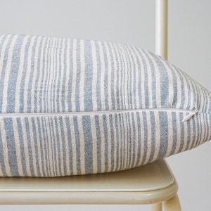 May include: A rectangular cushion with a blue and white striped design. The cushion features a zip closure on one side. The cushion is placed on a white chair.