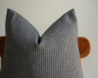 Puede incluir: Un cojín gris oscuro con rayas blancas verticales. El cojín está hecho de una tela texturizada y descansa sobre una silla de madera. El cojín tiene un diseño simple y minimalista, adecuado para la decoración del hogar.