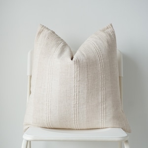 Puede incluir: Una almohada blanca con un sutil patrón de rayas se sienta en una silla blanca. La almohada está hecha de una tela suave y texturizada.