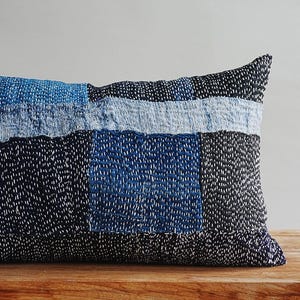 May include: A rectangular throw cushion featuring a patchwork design of indigo and black fabric panels. The surface is covered in dense, white running stitch embroidery, creating a textured appearance across the varied blue and dark fabric blocks.