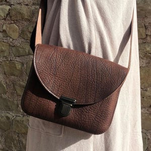 Puede incluir: Un bolso de cuero marrón cruzado con cierre de solapa y cierre de metal. El bolso tiene una superficie texturizada y una correa larga.