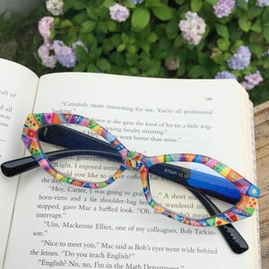 May include: Colorful patterned eyeglasses with black arms rest on an open book. The frames feature a vibrant mix of colors and geometric shapes. The text "R794P +2.50" is visible on the lens. The book is open to page 149.