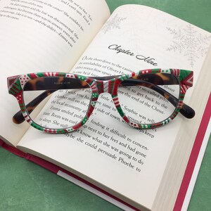 May include: Eyeglasses with a festive Christmas pattern in red, green, and white, resting on an open book. The glasses have a tortoiseshell design on the temples and a black frame. The book's title is "Chapter Nine."
