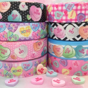 May include: Assortment of Valentine's Day themed ribbon rolls with candy heart designs. The ribbon rolls feature various patterns, including polka dots, stripes, and gingham. The candy hearts have messages like "Be Mine", "Hug Me", "Kiss Me", and "I Do".