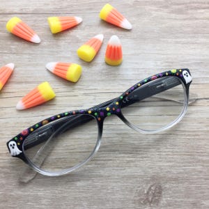 May include: Black reading glasses with a pattern of small white ghosts and colorful polka dots on the frames. The glasses feature clear lenses and are surrounded by several candy corn pieces, which are yellow, orange, and white triangular candies.