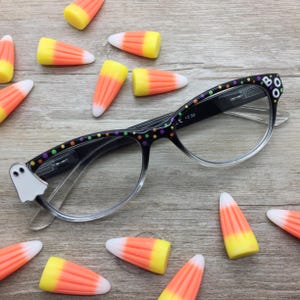 May include: Black reading glasses with clear lenses and a +2.50 magnification strength. The frames feature a pattern of colorful polka dots, a white ghost figure on the left temple, and the letters BOO on the right temple, surrounded by candy corn.