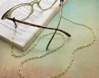 May include: Gold-colored eyeglasses chain draped over a pair of cat-eye glasses resting on an open book. The chain is made of rectangular links. The glasses have a light-colored frame and tortoise-shell arms.