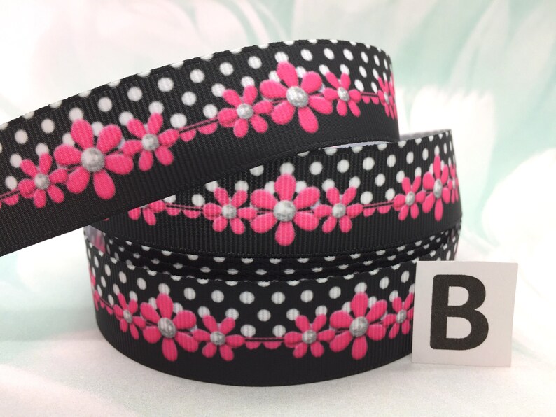 May include: Black and white polka dot ribbon with a row of pink flowers with silver centers. The ribbon is rolled up and the letter "B" is visible on a white label.