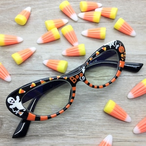 May include: Black cat-eye glasses with Halloween-themed decorations, including ghosts, pumpkins, and candy corn. The frames are black with orange and white accents, and the word "Boo" is featured on the frame. Candy corn candies are scattered around the glasses.