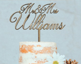 May include: Two-tiered wedding cake with white frosting and a rustic wood base. The cake is decorated with white daisies and gold pine cones. A wooden cake topper reads "Mr. & Mrs. Williams".