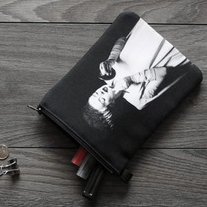 May include: Black and white zippered pouch featuring a vintage image of a woman. The pouch is open, revealing several pens and markers. A coin and a binder clip are next to the pouch on a gray wooden surface.
