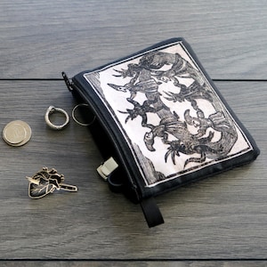 May include: Black zippered pouch with a detailed black and white illustration. Beside the pouch are two rings, two coins, and a heart-shaped pin with a knife through it. The items are on a gray wooden surface.