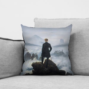 May include: A throw pillow featuring a painting of a man standing on a mountaintop overlooking a sea of clouds. The painting is in a muted color palette of grays, blues, and greens.