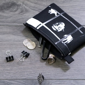 May include: A black and white zippered pouch with a graphic print of a person's face and hands. The pouch is open and contains coins, a small metal object, and a black binder clip. There are also three black binder clips on a gray wooden surface.