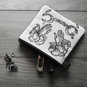 May include: A white zipped pouch featuring black illustrations of hands and tools. The pouch is open, revealing a utility knife and other items. Two binder clips and a coffee pot pin are next to the pouch on a grey wooden surface.