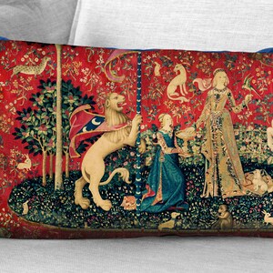 May include: A red, blue, and green tapestry pillow featuring a scene from the Unicorn Tapestries. A woman in a long gown stands between a lion and a unicorn, both of which are standing on their hind legs. The tapestry is surrounded by a border of flowers and leaves.