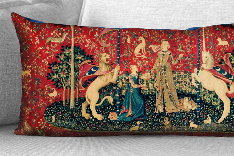 May include: A red, blue, and green tapestry pillow featuring a scene from the Unicorn Tapestries. A woman in a long gown stands between a lion and a unicorn, both of which are standing on their hind legs. The tapestry is surrounded by a border of flowers and leaves.