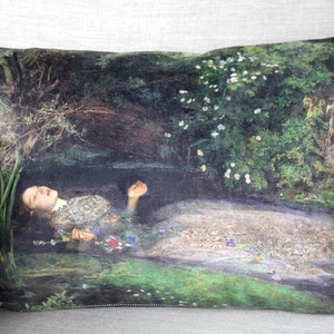 May include: A cushion featuring a printed image of a woman lying in a stream of water surrounded by flowers and greenery. The woman is wearing a white dress and has long, dark hair.