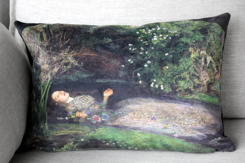 May include: A cushion featuring a printed image of a woman lying in a stream of water surrounded by flowers and greenery. The woman is wearing a white dress and has long, dark hair.