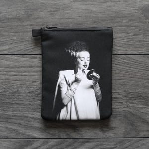 May include: Black rectangular zippered pouch featuring a black and white image of a woman applying makeup. The woman has dark hair and is wearing a white dress and gloves. The pouch is on a dark wood surface.