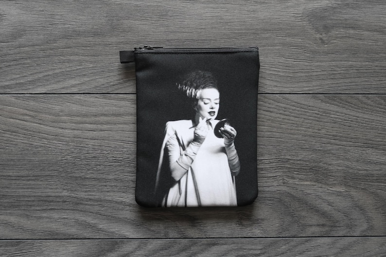 May include: Black rectangular zippered pouch featuring a black and white image of a woman applying makeup. The woman has dark hair and is wearing a white dress and gloves. The pouch is on a dark wood surface.