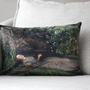 May include: A throw pillow with a black background featuring a painting of a woman lying in a forest stream. The woman is wearing a white dress and has long dark hair. The stream is surrounded by green foliage and white flowers.