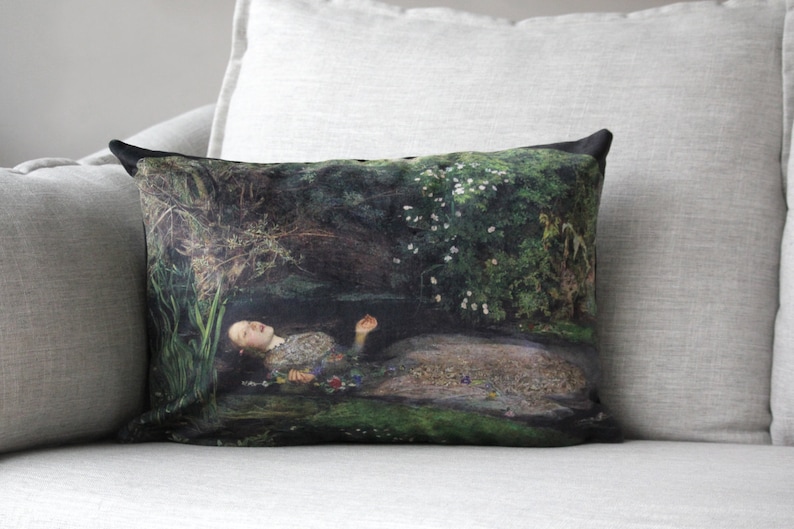 May include: A throw pillow with a black background featuring a painting of a woman lying in a forest stream. The woman is wearing a white dress and has long dark hair. The stream is surrounded by green foliage and white flowers.