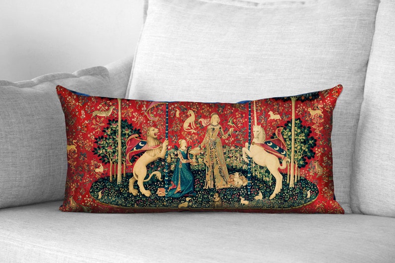May include: A red, blue, and green tapestry pillow featuring a unicorn and a lion with a woman in a blue dress and a woman in a gold dress. The tapestry is surrounded by a forest scene with trees, flowers, and animals.