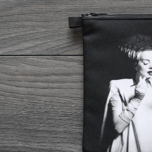 May include: Black zippered pouch featuring a black and white image of a woman in a white dress, holding a compact mirror. The image is a classic horror movie character. The pouch is on a gray wooden surface.